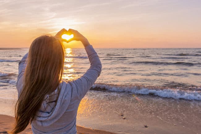 A woman standing on the beach holding her hands in the shape of a heart.